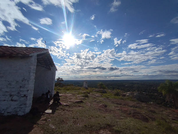 Bright sunlight shining over a small rural chapel on a hilltop, overlooking a vast natural landscape and forest horizon. The image conveys spirituality, solitude, travel, rural life, nature, faith, serenity, and scenic viewpoints in the countryside under a dramatic sky with clouds.
