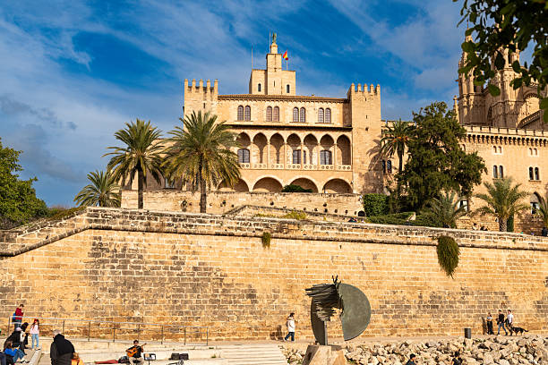 Palma - Balearic Islands - Spain - 12-28-2025: Royal Palace waterfront view shows a stone wall, arched palace facade, palm trees, and pedestrians beside a modern metal sculpture.