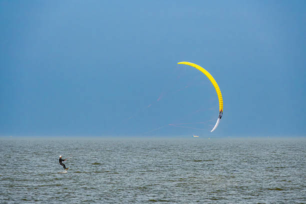 Amsterdam - North Holland - Netherlands - 04-11-2025: A kitesurfer rides over choppy water as a large yellow kite flies overhead, with taut lines and spray visible against bright blue sky.
