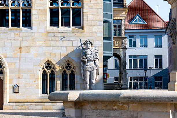 Quedlinburg - Saxony-Anhalt - Germany - 03-08-2025: Stone facade with arched windows and a carved knight statue, with modern buildings and a red tiled roof behind in bright sunlight.