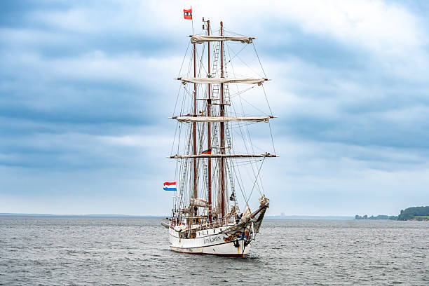 Groemitz - Schleswig-Holstein - Germany - 06-28-2025: Traditional tall sailing ship with furled sails moves across calm sea under cloudy sky, showing masts, rigging, and small flags aloft.