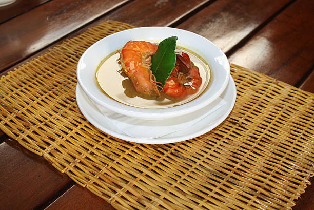 Cooked prawns presented in a white bowl with flavorful broth and a green leaf garnish, elegantly served on a woven placemat atop a wooden table