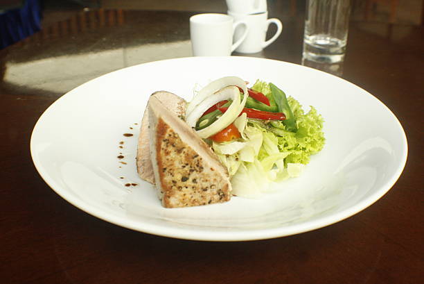 Delicious grilled tuna steak, perfectly seared and seasoned, served with a vibrant fresh green salad and balsamic drizzle on a white plate in a dining setting