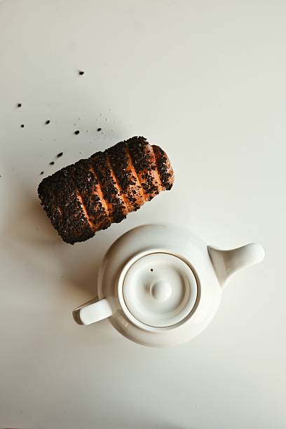 A single pastry, possibly a sweet roll or croissant, coated in dark crumbs sits beside a clean white teapot, suggesting a simple breakfast or snack setting
