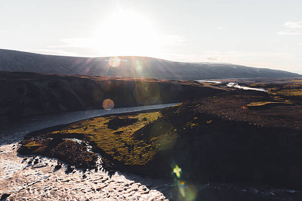 Wide angle view of an Icelandic river winding through a vast, rugged, and treeless arctic landscape, featuring a bright sun flare emphasizing the natural beauty and remote wilderness
