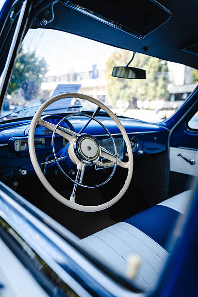 Close up of a classic car interior highlighting a vintage steering wheel, analog gauges, textured dashboard, and chrome details reflecting mid-century automotive design.