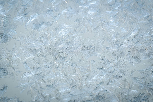 Light, feathery frost patterns spread gracefully across a pale glass surface, creating a soft and intricate winter texture. The icy formations resemble delicate brushstrokes or plumage, enhanced by gentle natural lighting. The overall tone is calm, minimal, and serene.