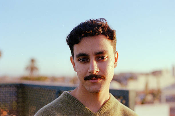 Young adult man smiling, wearing a sweater on a rooftop with urban background during sunny day. Scanned from analog film