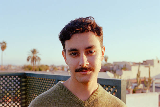 Young adult man with a mustache looking at the camera, standing on a city rooftop with palm trees in the background during golden hour. Scanned from analog film