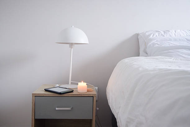 A minimalist bedside setup featuring a modern white lamp, a lit candle, and a digital tablet or e-reader on a wooden nightstand next to a realistic bed with white linen. Soft lighting creating a peaceful, hygge atmosphere