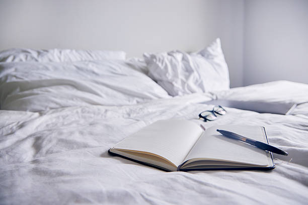 Cropped shot of young woman sits on bed, makes notes in diary, focused looking aside. Blogger create publication for blog in notebook. Copy space