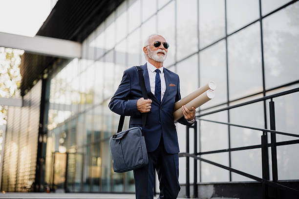 Older businessman in a suit carrying a laptop bag and rolled-up plans under his arm while exiting an office building. Corporate lifestyle, work routine, and professional business concept.