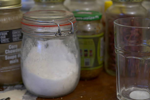 A horizontal close-up centers on a jar filled with a white substance, secured by a metal cage at its neck, with blurred jars behind and an empty glass in the foreground on a brown wooden table.