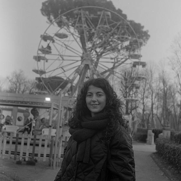 Young adult woman with curly hair smiling in front of a vintage amusement park with a ferris wheel. Scanned from analog film