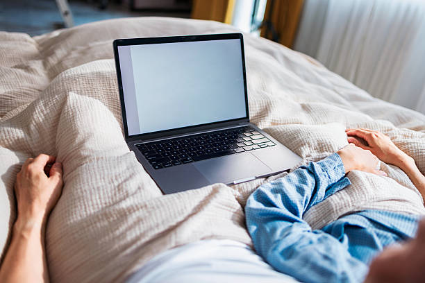 A person rests with a laptop on a soft bed under a beige blanket, conveying cozy work.
