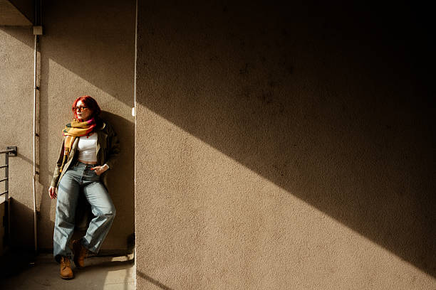 Young woman leaning against a wall in a narrow urban passage, dramatic sunlight and shadow creating a minimalist city lifestyle mood.