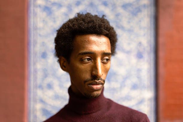 A portrait of a thoughtful man with curly hair, dressed in a maroon turtleneck, standing before a beautifully patterned blue and white wall.