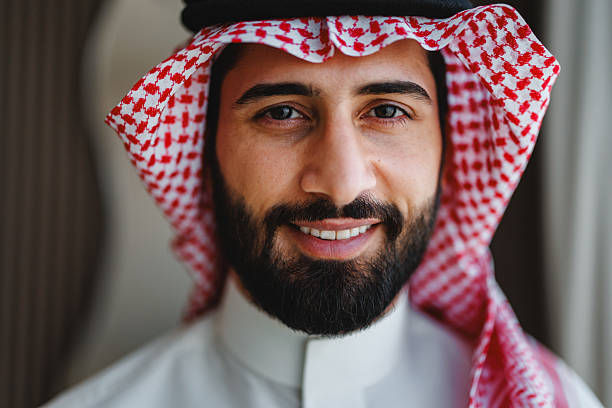 Portrait of a young Saudi man in traditional clothing, featuring a cheerful smile and a bright expression. He wears a red and white checkered headscarf and a white robe, creating a classic and cultural appearance.