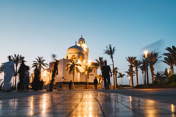 People walking along a promenade with palm trees near an illuminated mosque. The sky is clear and transitioning from day to night, creating a serene atmosphere. Lights from the mosque and walkway cast reflections on the tiled path.