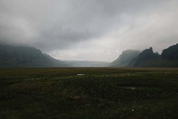Expansive green valley with distant mountains shrouded in low-hanging clouds and a moody, atmospheric sky creating a dramatic natural scene in Iceland