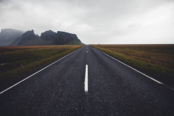 Empty asphalt road stretches towards the horizon, framed by flat, barren fields and dark, rugged mountains under an overcast sky in the remote Icelandic countryside