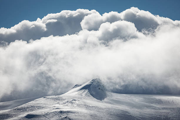 Snow-capped mountain peak piercing low, thick clouds beneath a clear blue sky, evoking Iceland’s remote, tranquil arctic winter landscape and untouched high-elevation solitude