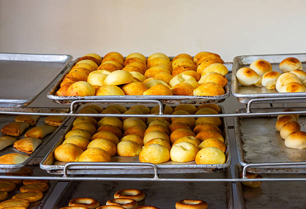 Stacks of golden bread rolls arranged neatly on metal trays, showcasing a variety of shapes and sizes in a warm setting.