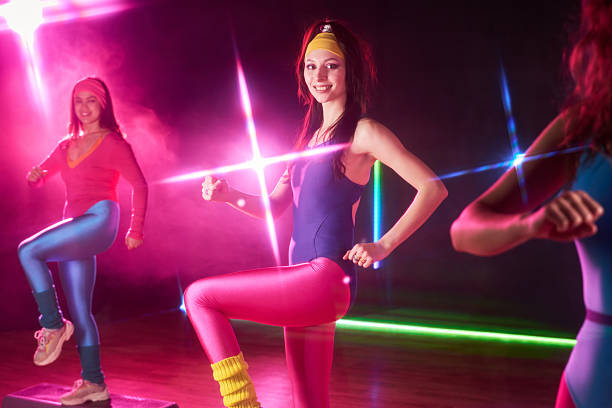 Group of young adult Caucasian women exercising in fitness studio, performing aerobic step workout in colorful activewear, smiling and looking forward under bright stage lights