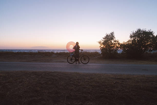 Photo of a cheerful mid adult woman, biking home after day spent on local beach while holding inflatable swim ring