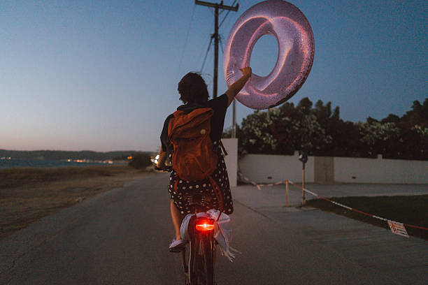 Photo of a cheerful mid adult woman, biking home after day spent on local beach while holding inflatable swim ring