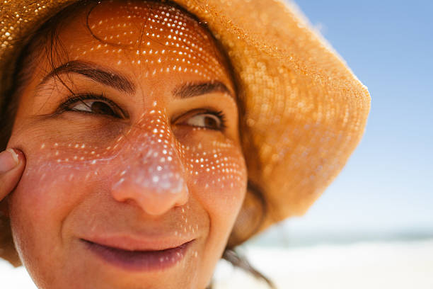 Photo of a mid adult woman applying sunscreen on her face while at the beach