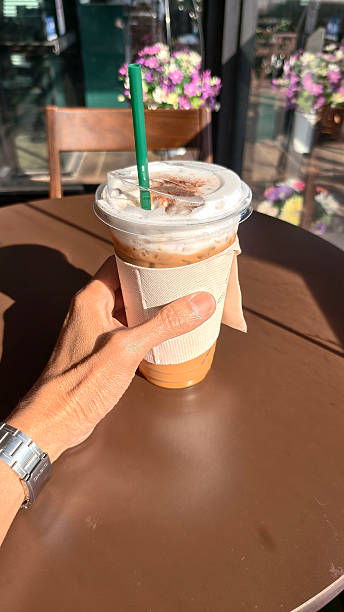 POV hand holding an iced coffee with foam and green straw at sunlit cafe table. Bright morning light creates soft shadows on the brown tabletop in cafe and purple flowers