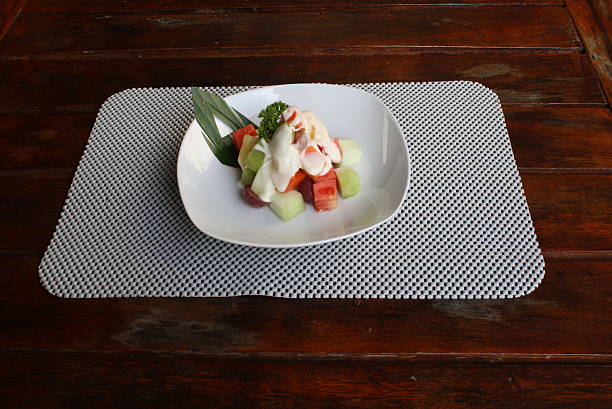 Colorful fruit salad with diced watermelon, melon, and grapes, topped with creamy dressing and parsley, served on a white plate on a dark wooden table