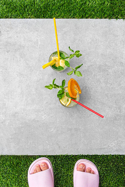 Two colorful citrus drinks with orange slices and mint garnish rest on a patio surface bordered with lush green grass. Pink flip-flops add a summer vibe, suggesting relaxation and freshness
