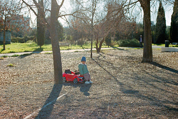 Young child wearing a blue hat and green jacket sitting on a red toy car, playing outdoors in a sunny park. Scanned from analog film