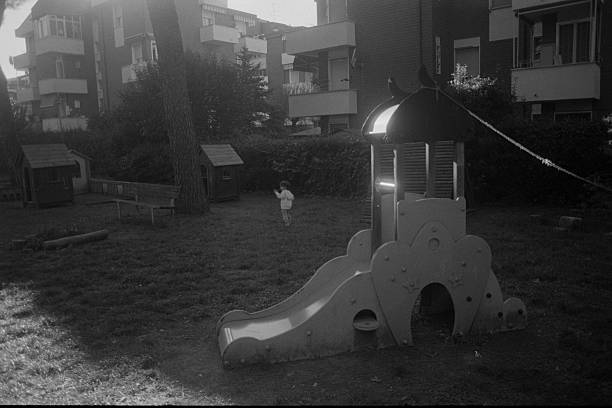 Young child walking alone in a public park playground with slide and playhouses, black and white film aesthetic. Scanned from analog film