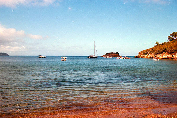 Serene view of a sailboat and leisure boats anchored near a tropical island coast, reflecting a peaceful summer vacation. Scanned from analog film