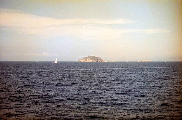 Sailboat smoothly navigating a calm blue sea near a small rocky island under a bright sky, symbolizing freedom and adventure. Scanned from analog film