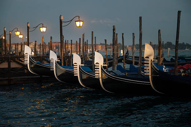 A row of traditional gondolas rests along wooden mooring poles in Venice, gently floating on the lagoon at dusk. Soft evening light illuminates the iconic curved prows and iron ferro details, while warm reflections shimmer across the water. The scene captures the charming and timeless atmosphere of Venice, highlighting its historic boats, tranquil waterfront, and romantic maritime character.
