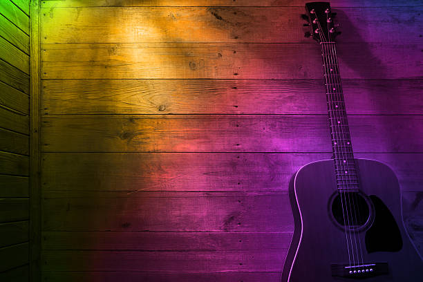 The acoustic guitar is leaning against a wooden wall lit by a multi colored light, creating a cozy atmosphere. Space for copy.
