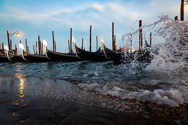 Traditional gondolas are moored along the waterfront in Venice, with waves splashing against the stone edge of the lagoon. The scene captures the relationship between the historic boats and the surrounding water, highlighting movement, reflections, and the maritime atmosphere of the Venetian lagoon at dusk.