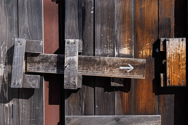 A handmade wooden sliding latch on a weathered wooden door. A white arrow indicating the direction of movement is painted on the bolt. A close-up with a retro and rustic atmosphere.
