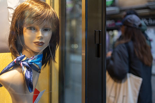 Madrid. Spain. Mannequin head, wearing a brown wig and a blue patterned scarf, stands in a shop display window, reflecting a blurred street scene with pedestrians walking by