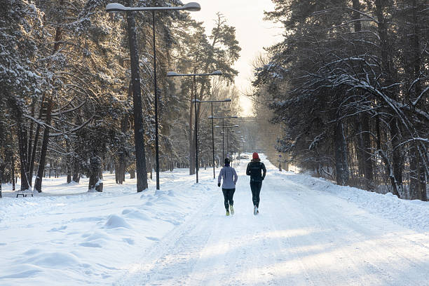 Two people jogging along a snow-covered forest road in winter, exercising outdoors in cold weather conditions.