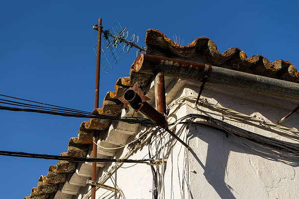 A weathered and rusty analog TV antenna mounted on a traditional tiled roof. Multiple messy electrical and telecommunication cables hang against a white-washed wall under a clear blue sky. Concept of outdated technology, rural infrastructure, and poor utility management.