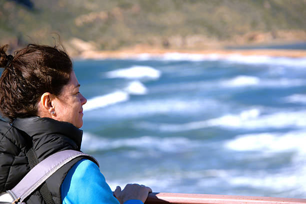 A woman at a lookout point observes the waves of a choppy sea on a sunny winter day.