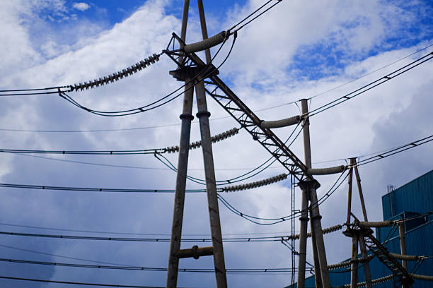 A network of high voltage power lines and sturdy insulators extends across the frame set against a dynamic blue sky with scattered clouds indicating power generation or transmission