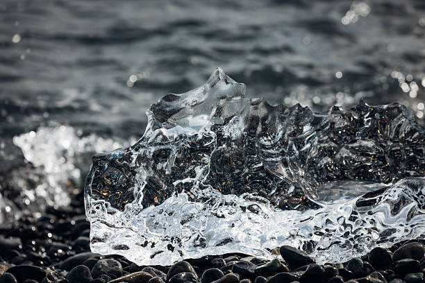 Iceberg fragment with a unique shape sparkling on the black volcanic sand beach, with ocean waves creating a natural and dynamic scene capturing the cold beauty of Iceland's glacial landscapes