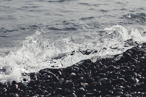 Sparkling blue water splashes against a collection of dark volcanic pebbles, highlighting the rugged natural beauty and powerful forces of the Icelandic coast