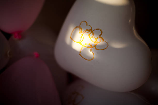 Romantic wedding decoration featuring elegant silver foil heart-shaped balloons with white ribbons floating in a sophisticated reception hall. The balloons are arranged against soft pink walls with cream-colored curtains, creating an ethereal and romantic atmosphere. The venue's classic interior with its warm lighting and traditional artwork on the walls adds a timeless elegance to the celebration setup.